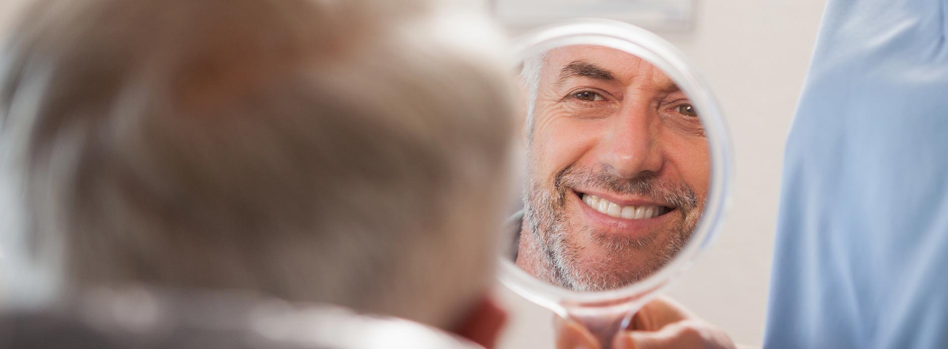 The image shows a man with a beard smiling at his reflection in a mirror while sitting in a barber s chair, with another person visible in the background.
