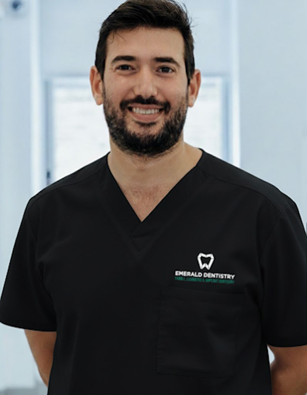 The image shows a man wearing a white coat with a name tag, standing indoors with a smile on his face.